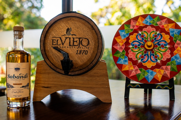 A tequila barrel, colorful mandala plate, and a bottle on a wooden table.