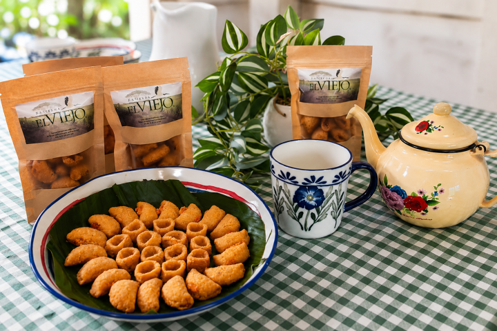 Cookies on a plate, teapot, mugs, plant, and packaged snacks on a checkered tablecloth.