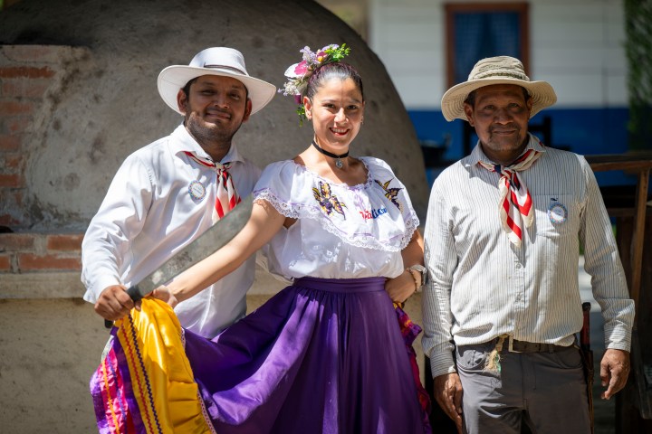 Three people in traditional attire smiling, with a woman in a purple skirt between two men.