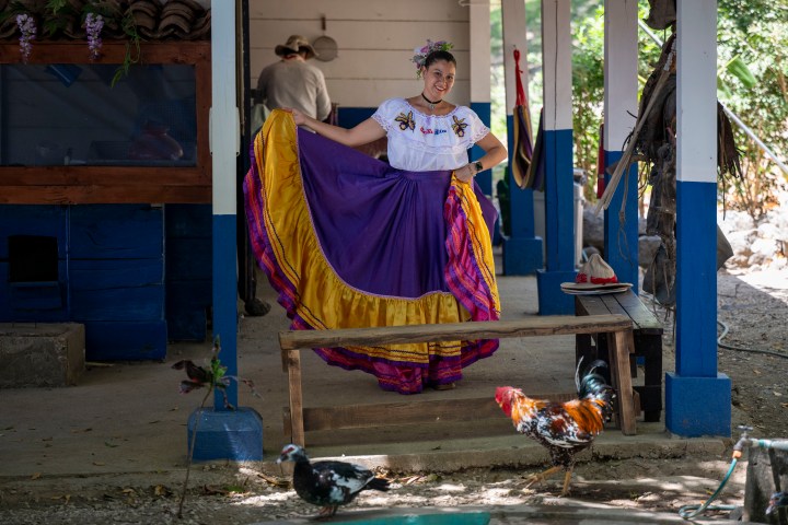 Woman in colorful dress posing near bench, with chickens nearby.