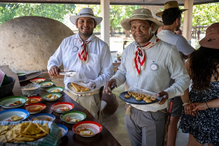 Two men in hats serving food at an outdoor gathering with colorful plates on a table.