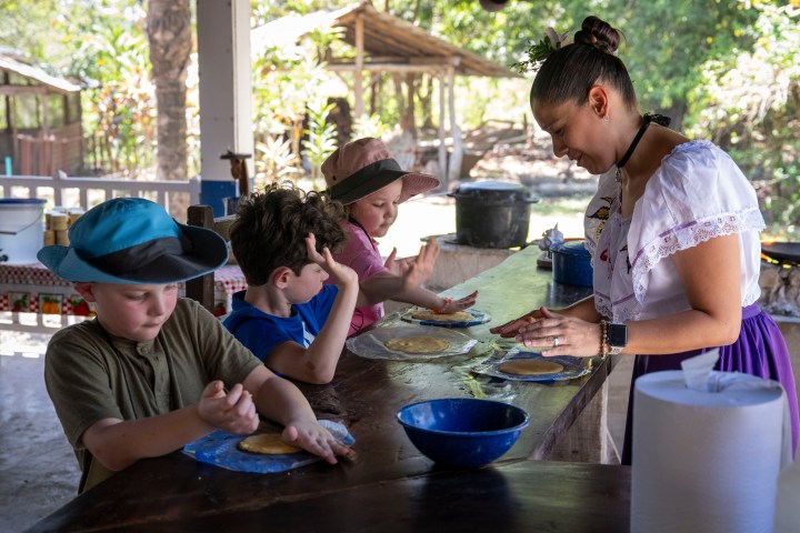 Children and woman making dough at outdoor table with trees in background.