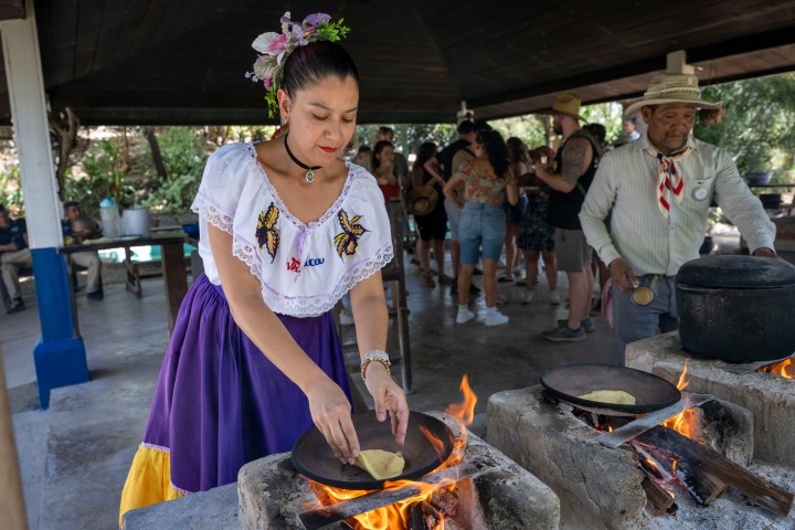 Woman in traditional dress cooking outdoors on an open fire, with a group of people in the background.