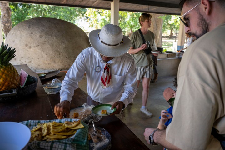 Person in hat serves food at an outdoor table with onlookers.