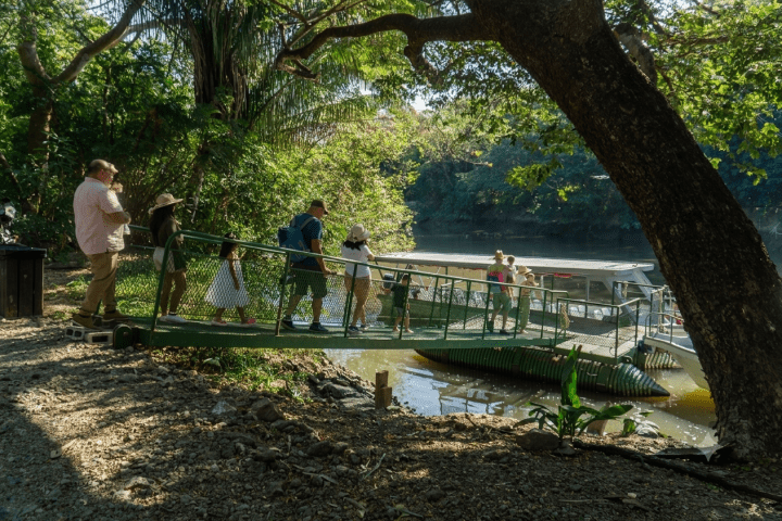 People board a boat on a river surrounded by lush trees.