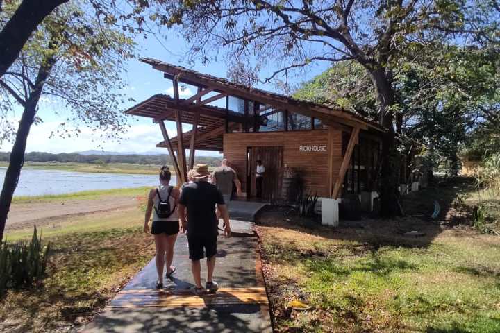 People walking towards a wooden building by a lake under tree shade.