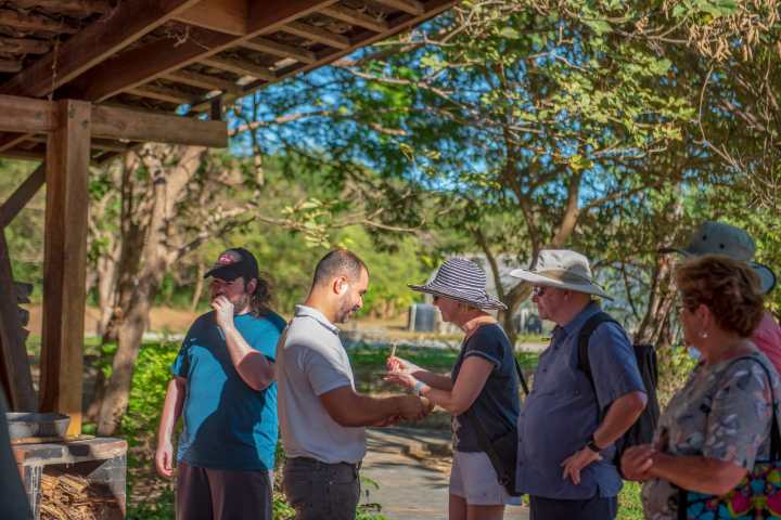 Group of people under a wooden canopy in a sunny outdoor setting, some wearing hats.