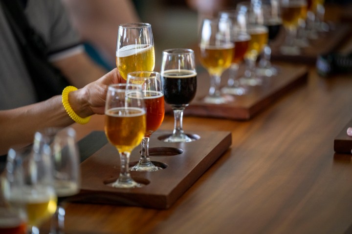 Person holding a glass from a flight of various beers on a wooden tray.