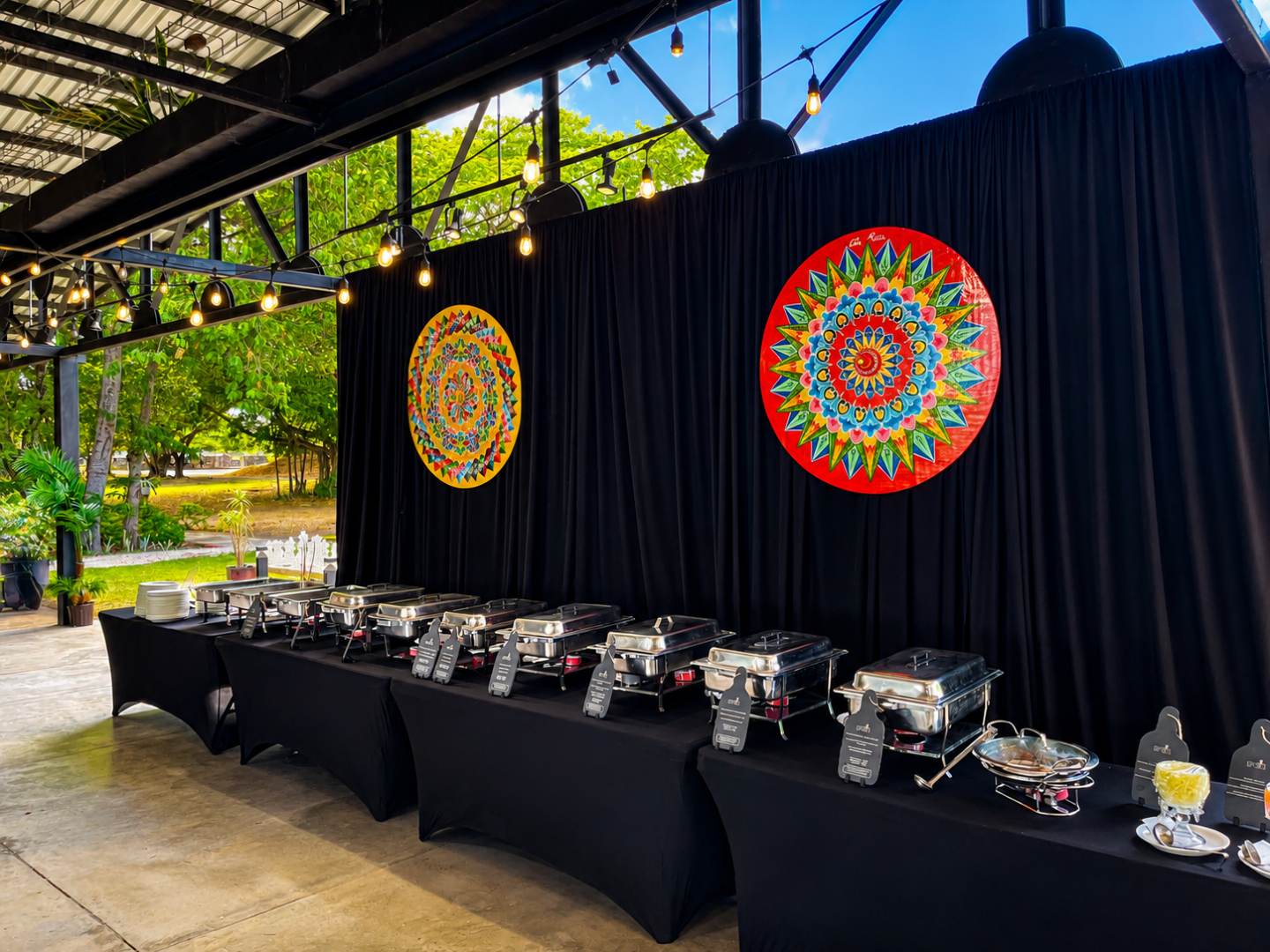 Buffet setup with covered chafing dishes and colorful circular art on black drapes.