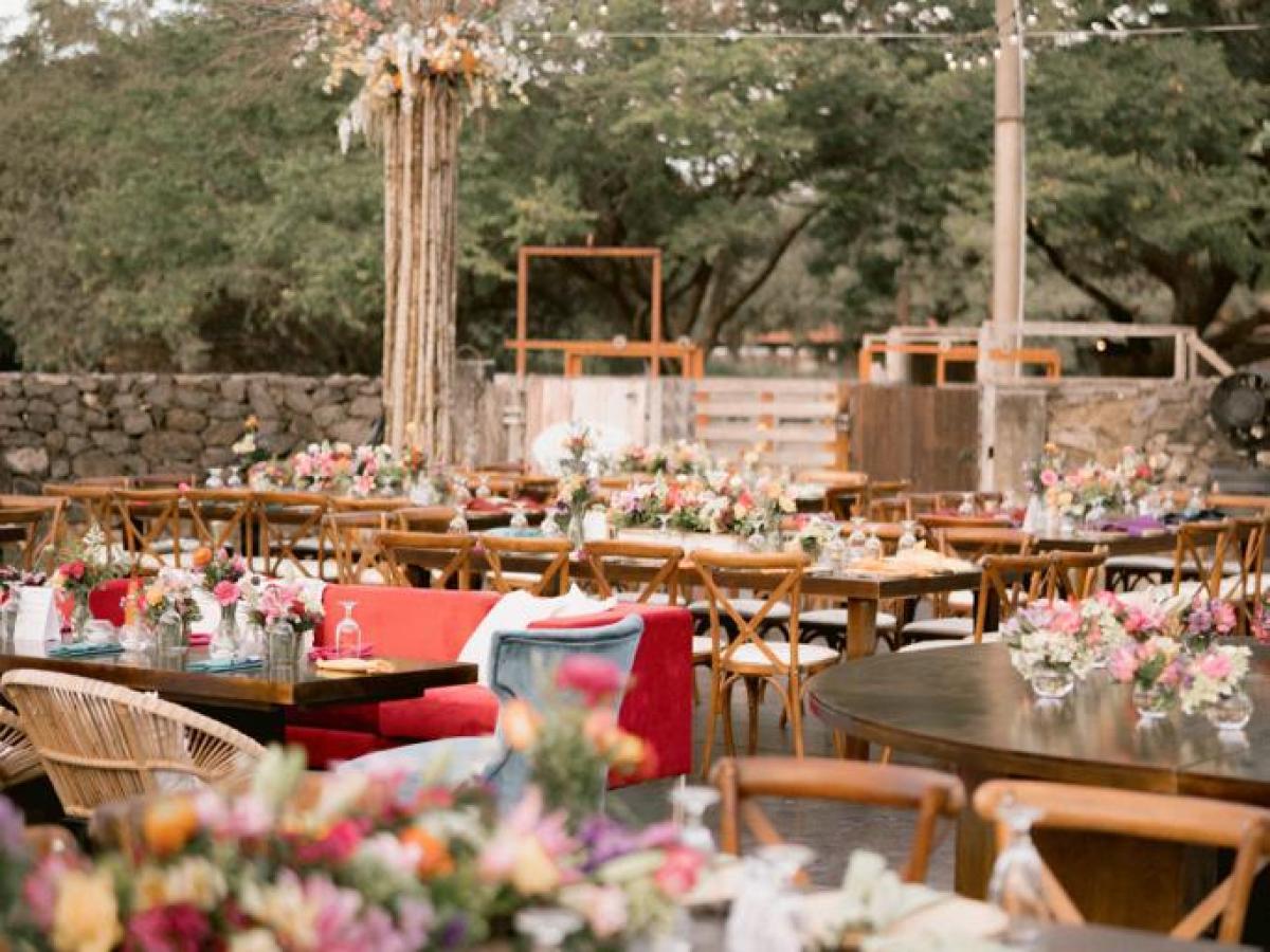 Outdoor event with tables set, floral centerpieces, string lights, surrounded by trees.