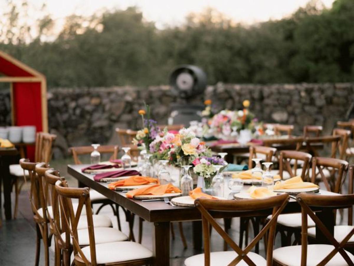 Outdoor dining setup with wooden tables and chairs, decorated with colorful flowers and napkins.